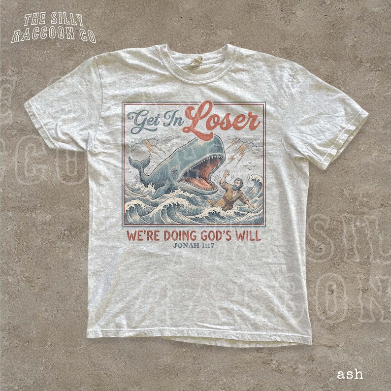 A white t-shirt with a graphic design depicting a person being attacked by a large whale, with the text "Get In Loser" and "We're Doing God's Will" below the image.