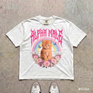 A white t-shirt with a graphic design featuring an orange cat surrounded by flowers and a rainbow.