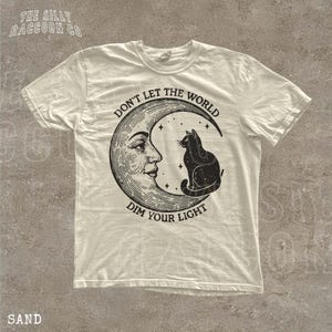 A white t-shirt with a graphic design depicting a cat sitting on a crescent moon and the text "Don't let the world dim your light" printed on it.