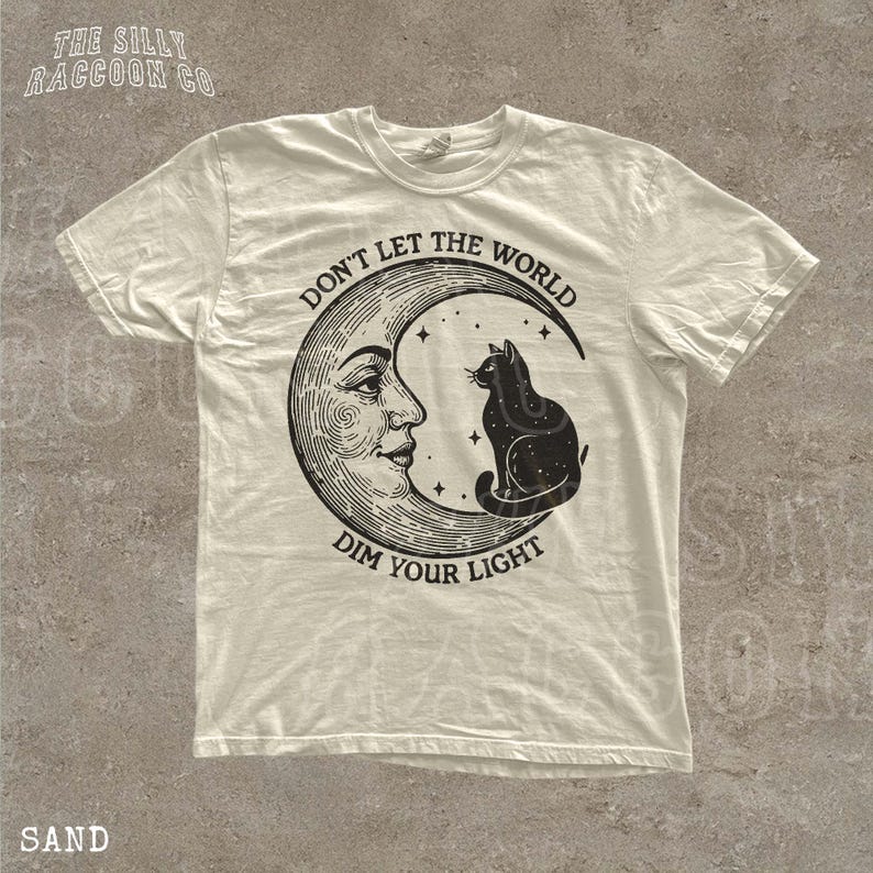 A white t-shirt with a graphic design depicting a cat sitting on a crescent moon and the text "Don't let the world dim your light" printed on it.