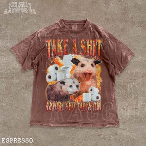 A t-shirt with a graphic design featuring an opossum surrounded by toilet paper rolls, with the text "TAKE A SHIT BEFORE SHIT TAKES YOU" prominently displayed.