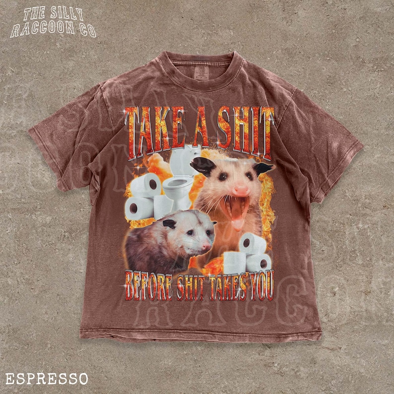 A t-shirt with a graphic design featuring an opossum surrounded by toilet paper rolls, with the text "TAKE A SHIT BEFORE SHIT TAKES YOU" prominently displayed.