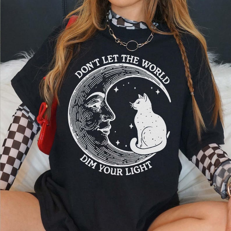 A person wearing a black t-shirt with a graphic design of a cat on a crescent moon and the text "Don't Let the World Dim Your Light".