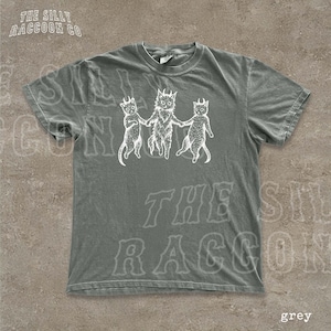 A grey t-shirt with a graphic design featuring three cats with horns and a cat with a lion's head, all holding hands.