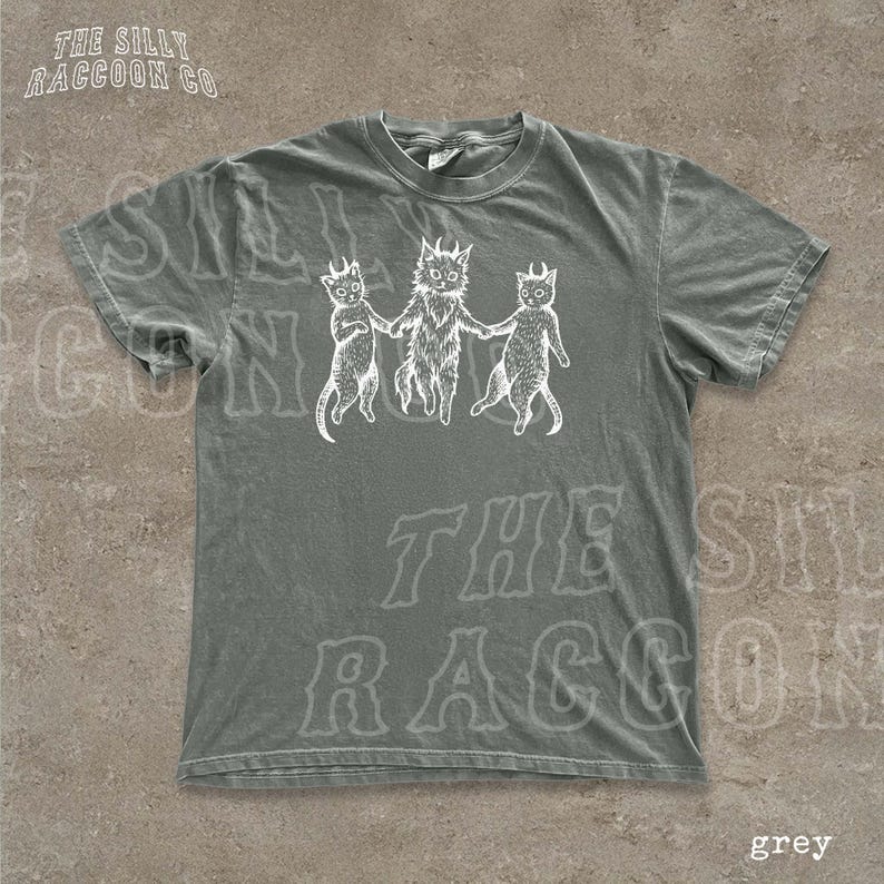 A grey t-shirt with a graphic design featuring three cats with horns and a cat with a lion's head, all holding hands.