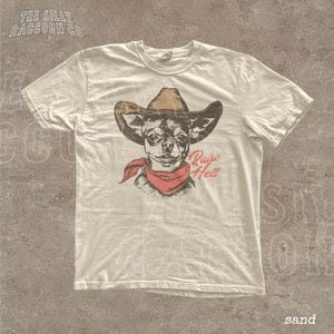 A white t-shirt with a graphic design of a dog wearing a cowboy hat and a red bandana, along with the text "Raise Hell" printed on it.