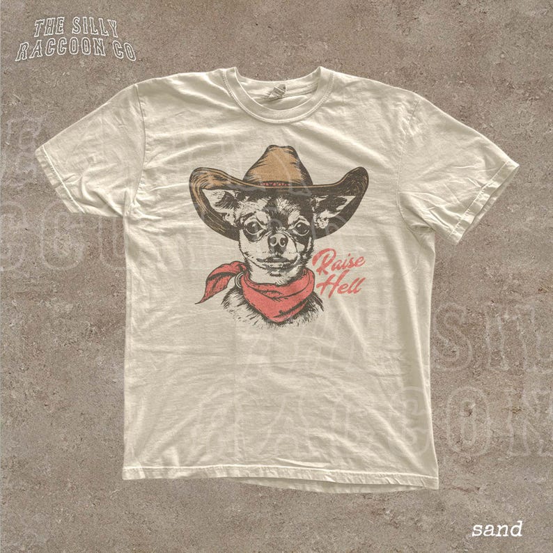 A white t-shirt with a graphic design of a dog wearing a cowboy hat and a red bandana, along with the text "Raise Hell" printed on it.