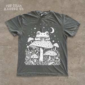 May include: Dark gray t-shirt with a white graphic of a frog sitting on a mushroom, surrounded by other mushrooms, stars, and a crescent moon. The text "THE SILLY RACCOON CO" is printed at the top left.