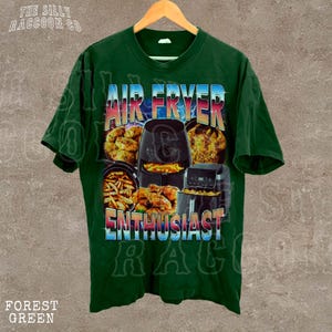 The image is a green t-shirt with a graphic design featuring an air fryer and various fried food items, along with the text "AIR FRYER ENTHUSIAST" and "THE SILKY RACCOON CO."