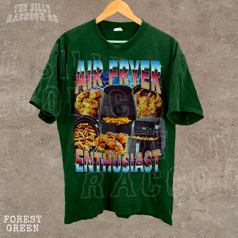 The image is a green t-shirt with a graphic design featuring an air fryer and various fried food items, along with the text "AIR FRYER ENTHUSIAST" and "THE SILKY RACCOON CO."