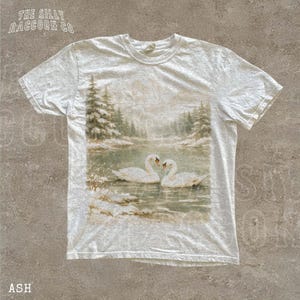 A white t-shirt with a printed image of two swans swimming in a lake surrounded by snow-covered trees.