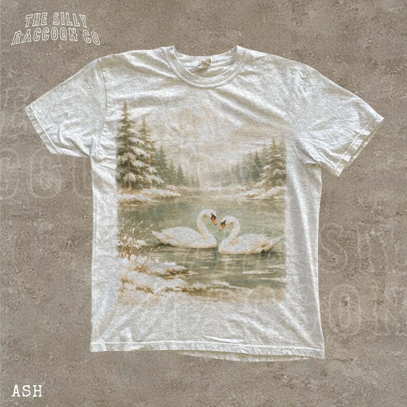 A white t-shirt with a printed image of two swans swimming in a lake surrounded by snow-covered trees.