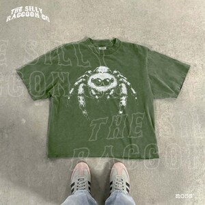 A pair of white sneakers and a green t-shirt with a spider graphic are displayed on a concrete floor.