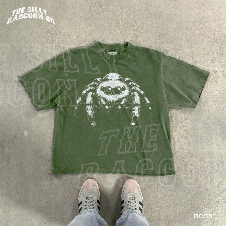 A pair of white sneakers and a green t-shirt with a spider graphic are displayed on a concrete floor.