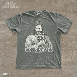 A gray t-shirt with a graphic design depicting a man holding a raccoon. The text below the graphic says "JESUS SAVES" and "LUKELINE 1910".