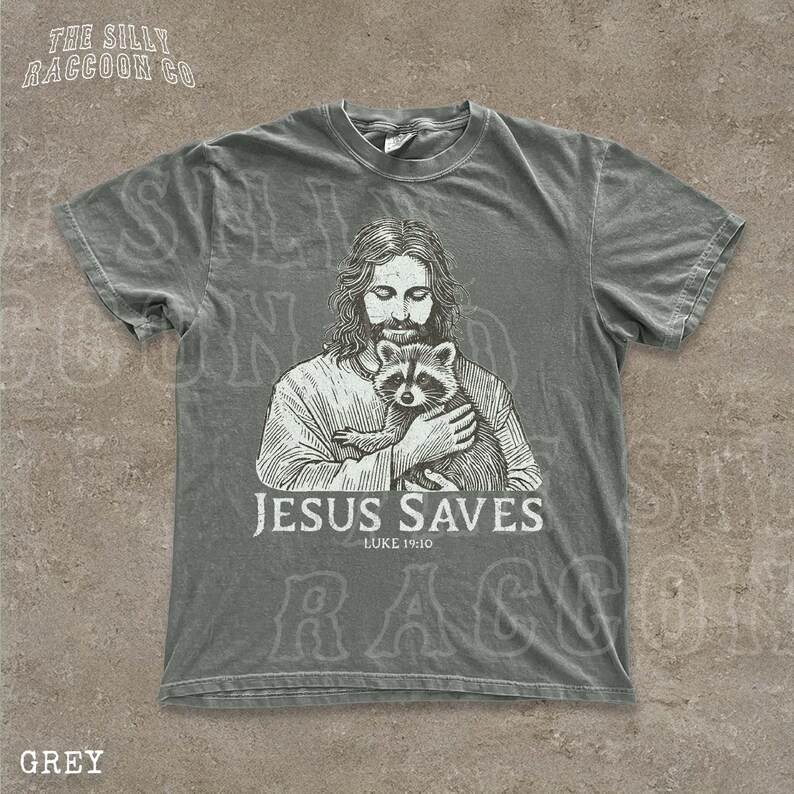 A gray t-shirt with a graphic design depicting a man holding a raccoon. The text below the graphic says "JESUS SAVES" and "LUKELINE 1910".