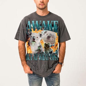 A man wearing a gray t-shirt with a graphic design featuring a koala and the text "A WAKE BUT AT WHAT COST".