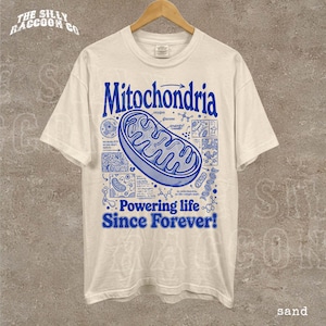 A white t-shirt with a graphic design depicting Mitochondria, a cellular organelle, and text that reads "Mitochondria Powering life Since Forever!"