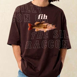 A man wearing a brown t-shirt with a fish graphic and the word "fih" printed on it.