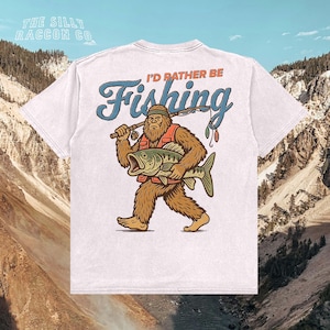 A white t-shirt with a graphic design depicting a large, furry creature holding a fish and the text "I'D Rather Be Fishing" printed on it.