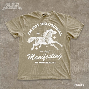 a t-shirt with a graphic design of a horse and the text "I'm not delusional, I'm just Manifesting, MY OWN REALITY" printed on it.