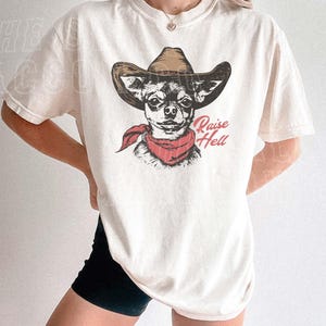 A woman wearing a white t-shirt with a graphic of a dog wearing a cowboy hat and a red scarf.