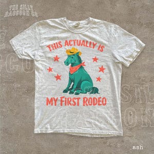 a white t-shirt with a graphic design depicting a green cartoon horse wearing a cowboy hat and a red bandana, surrounded by red stars. The text on the t-shirt reads "This actually is my first rodeo", and there is a small text "ash" at the bottom right corner.