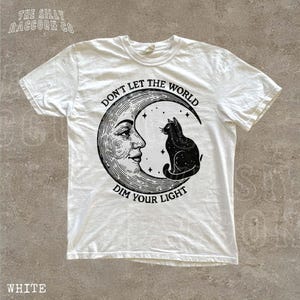 A white t-shirt with a graphic design featuring a cat sitting on a crescent moon and the text "Don't let the world dim your light" printed on it.