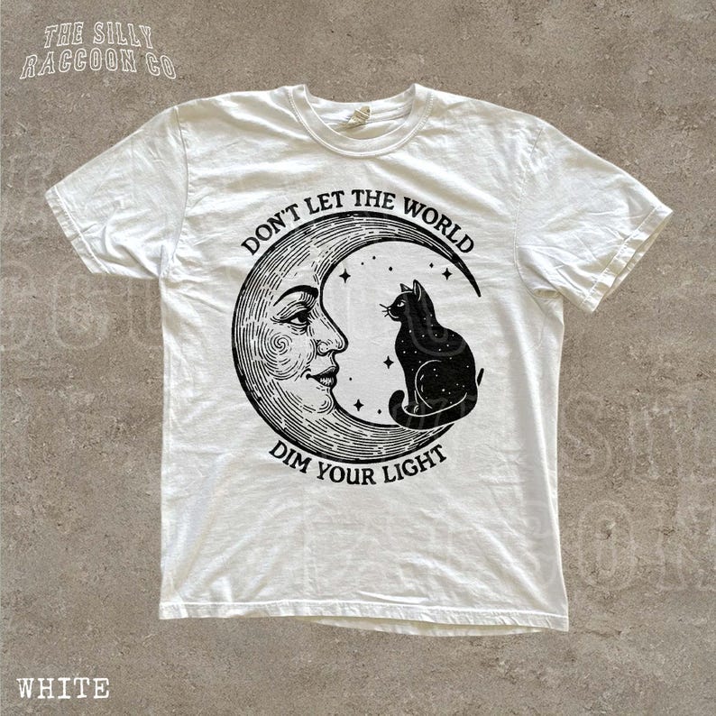 A white t-shirt with a graphic design featuring a cat sitting on a crescent moon and the text "Don't let the world dim your light" printed on it.
