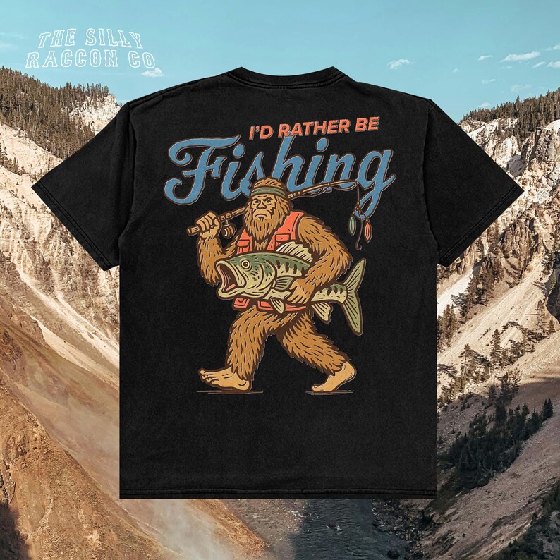 A black t-shirt with a graphic design depicting a large, furry creature holding a fishing rod and a fish, with the text "I'D Rather Be Fishing" printed on the back.