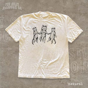 A white t-shirt with a graphic design featuring three cats with horns, depicted in a whimsical and stylized manner.