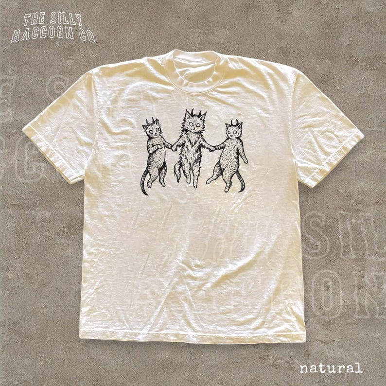 A white t-shirt with a graphic design featuring three cats with horns, depicted in a whimsical and stylized manner.