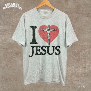 May include: A light gray t-shirt with the text "I Jesus" in black, with a red heart featuring a depiction of Jesus on the cross. The shirt is hanging on a wooden hanger.