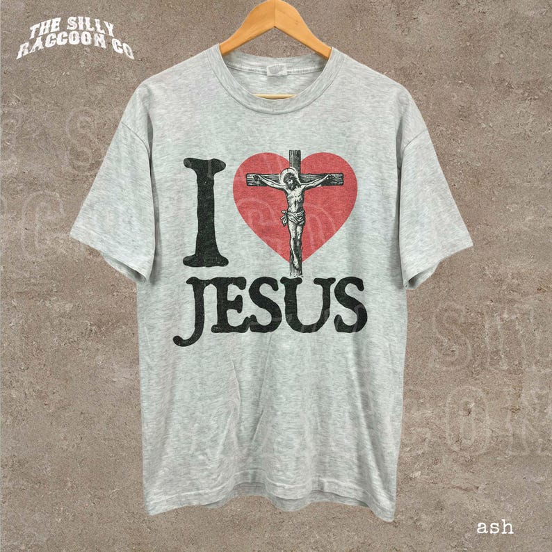 May include: A light gray t-shirt with the text "I Jesus" in black, with a red heart featuring a depiction of Jesus on the cross. The shirt is hanging on a wooden hanger.