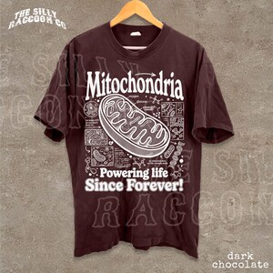 A dark brown t-shirt with a graphic design depicting Mitochondria, a cellular organelle, and text that reads "Mitochondria. Powering life since forever!"