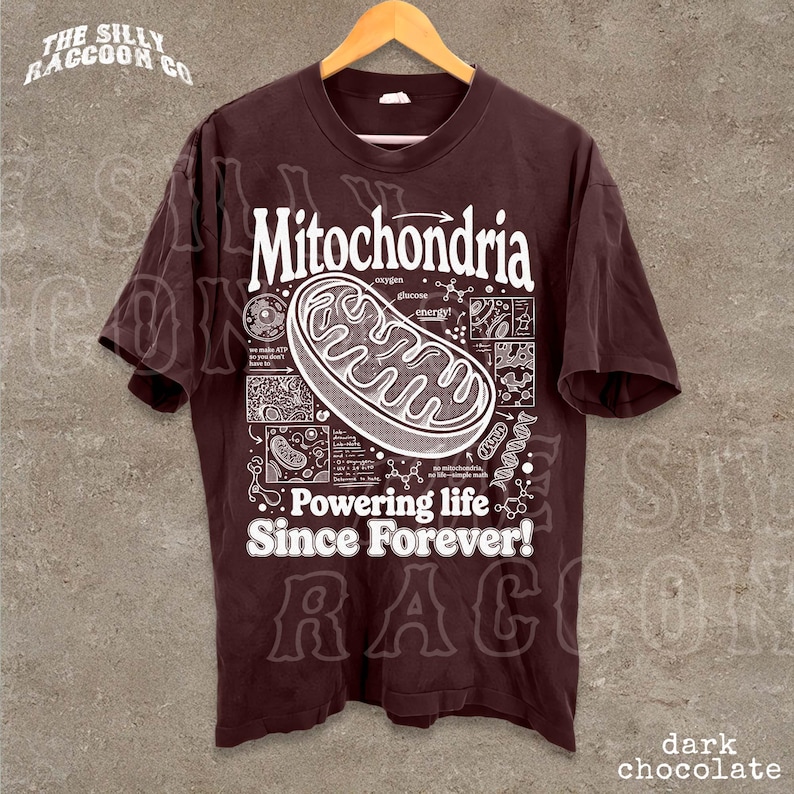 A dark brown t-shirt with a graphic design depicting Mitochondria, a cellular organelle, and text that reads "Mitochondria. Powering life since forever!"