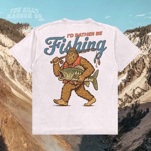 A white t-shirt with a graphic design depicting a large, furry creature holding a fish and the text "I'D Rather Be Fishing" printed on it.