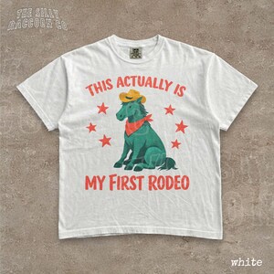 A white t-shirt with a graphic design featuring a green cartoon horse wearing a cowboy hat and a red bandana, surrounded by red stars. The text on the t-shirt reads "THIS ACTUALLY IS MY FIRST RODEO".