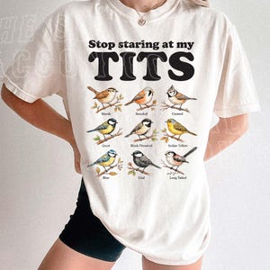 A person wearing a white t-shirt with a graphic design of various bird species and the text "Stop staring at my TITS" printed on it.