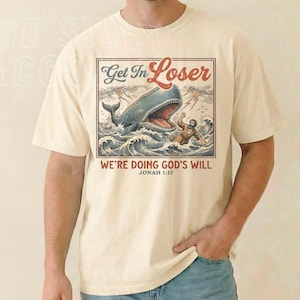 A man wearing a white t-shirt with a graphic design depicting a whale and a person, along with the text "Get In Loser" and "WE'RE DOING GOD'S WILL" printed on it.