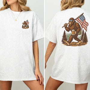 K&ouml;nnte beinhalten: Hellgraues T-Shirt mit einem Bigfoot-Motiv und einer amerikanischen Flagge. Auf der Vorderseite befindet sich ein kleines Bigfoot-Design, w&auml;hrend die R&uuml;ckseite eine gr&ouml;&szlig;ere Illustration von Bigfoot mit einer Flagge und einem Bier zeigt. Das T-Shirt besteht aus einem weichen, bequemen Material.