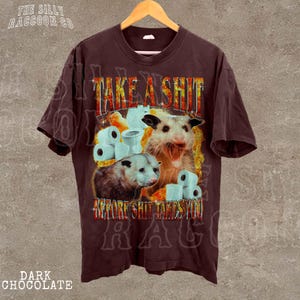 A t-shirt with a graphic design featuring two opossums and the text "TAKE A SHIT BEFORE SHIT TAKES YOU".