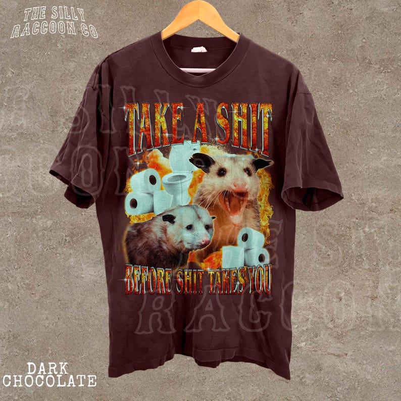 A t-shirt with a graphic design featuring two opossums and the text "TAKE A SHIT BEFORE SHIT TAKES YOU".