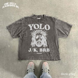 A person's feet wearing sneakers are standing on a concrete floor, with a gray t-shirt featuring a graphic design and text printed on it.