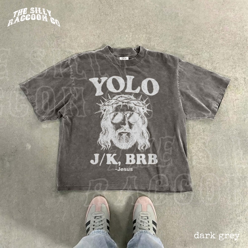 A person's feet wearing sneakers are standing on a concrete floor, with a gray t-shirt featuring a graphic design and text printed on it.