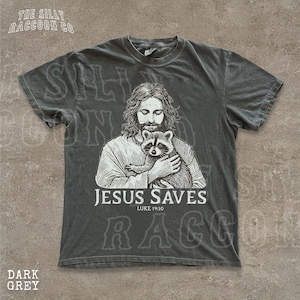 A t-shirt with a graphic design depicting a man holding a raccoon, along with the text "JESUS SAVES" and "LUKELI 19:10".