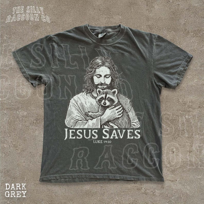 A t-shirt with a graphic design depicting a man holding a raccoon, along with the text "JESUS SAVES" and "LUKELI 19:10".