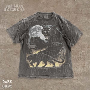 Puede incluir: Camiseta gris oscuro con un diseño de panteras negras y una luna llena. La camiseta tiene un aspecto vintage y desgastado y presenta el texto "THE SILLY RACCOON CO" en la parte superior.
