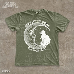 A green t-shirt with a white illustration of a cat sitting on a crescent moon and the text "Don't Let the World Dim Your Light" printed on it.