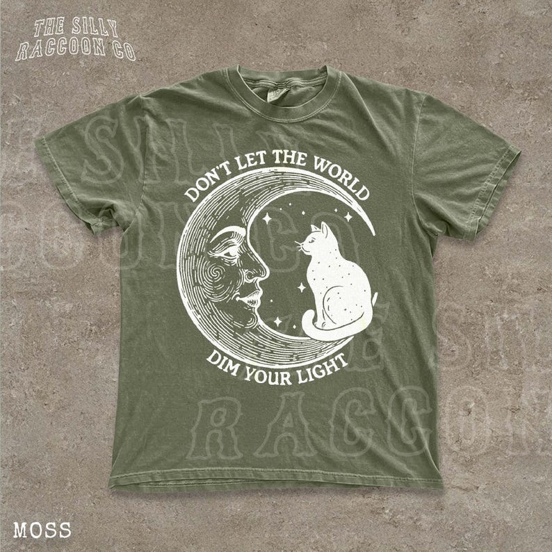 A green t-shirt with a white illustration of a cat sitting on a crescent moon and the text "Don't Let the World Dim Your Light" printed on it.